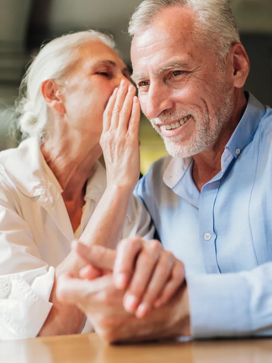 Oudere vrouw fluistert glimlachend iets in het oor van een oudere man